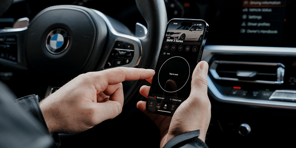 Person using a smartphone with an OBDeleven diagnostic app to scan BMW, with the steering wheel and dashboard in view.