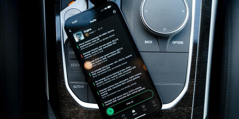 Smartphone screen displaying an OBDeleven diagnostic app with a list of engine faults, placed on the center console of a car next to the gear shift and control dials.