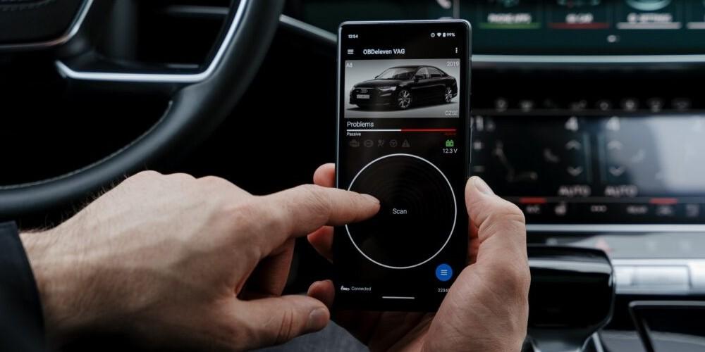  A person’s hands holding a smartphone with the OBDeleven app open, displaying diagnostic data for a car, with the interior of the vehicle and car dashboard visible in the background.
