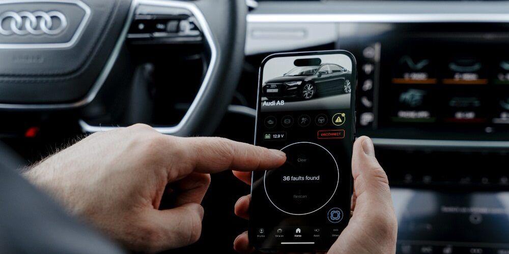  person's hands holding a smartphone with an OBDeleven diagnostic app displaying "36 faults found" in a car, with the interior and dashboard of an Audi visible in the background.